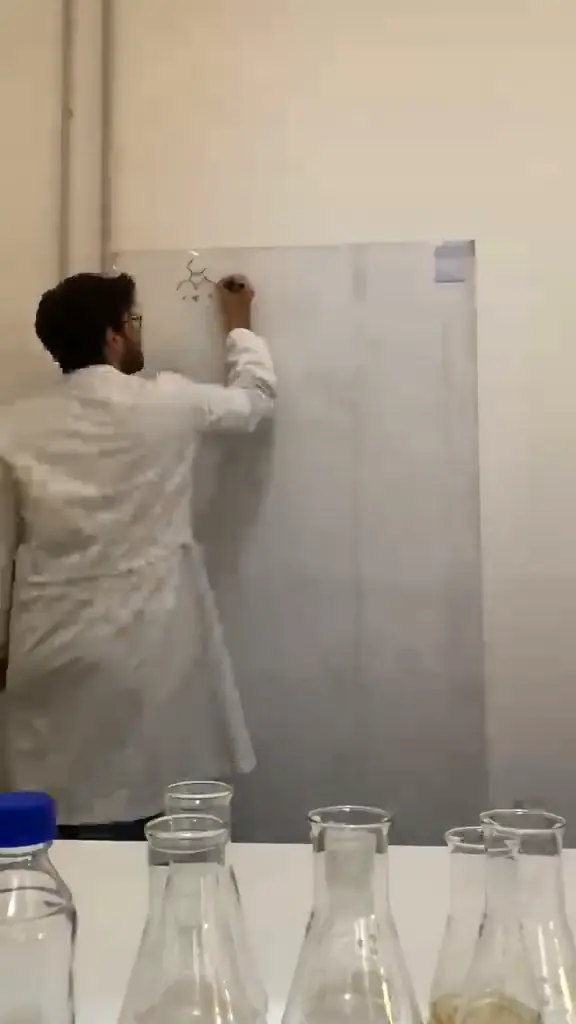 Uno studente in camice disegna una molecola alla lavagna, con le beute in primo piano
